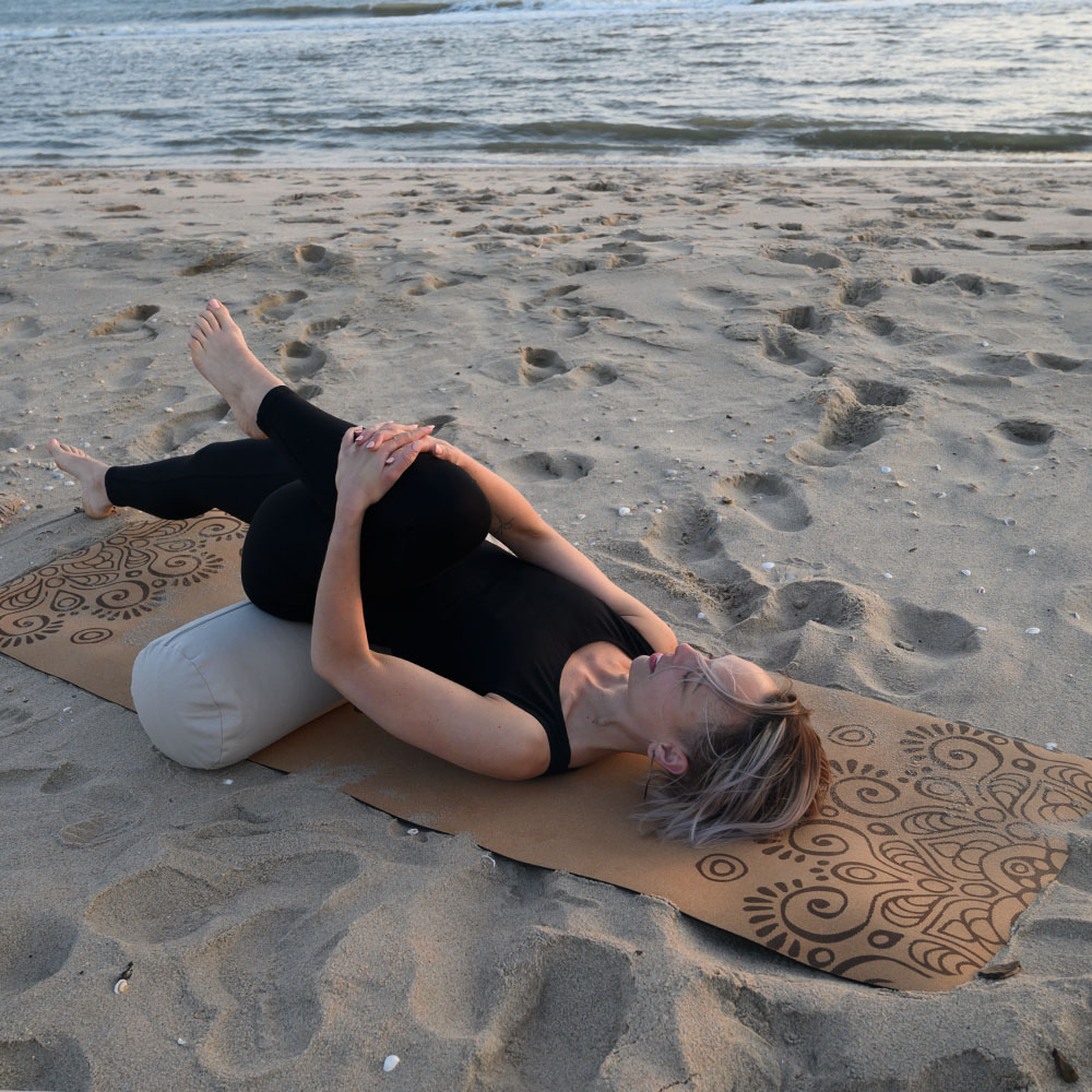 Jumpsuit Oceana Zwart #3 Een vrouw die een houding aanneemt in een herstellende asana, waarbij ze een bolster als steun gebruikt en op de yogamat van kurk en rubber op het strand ligt